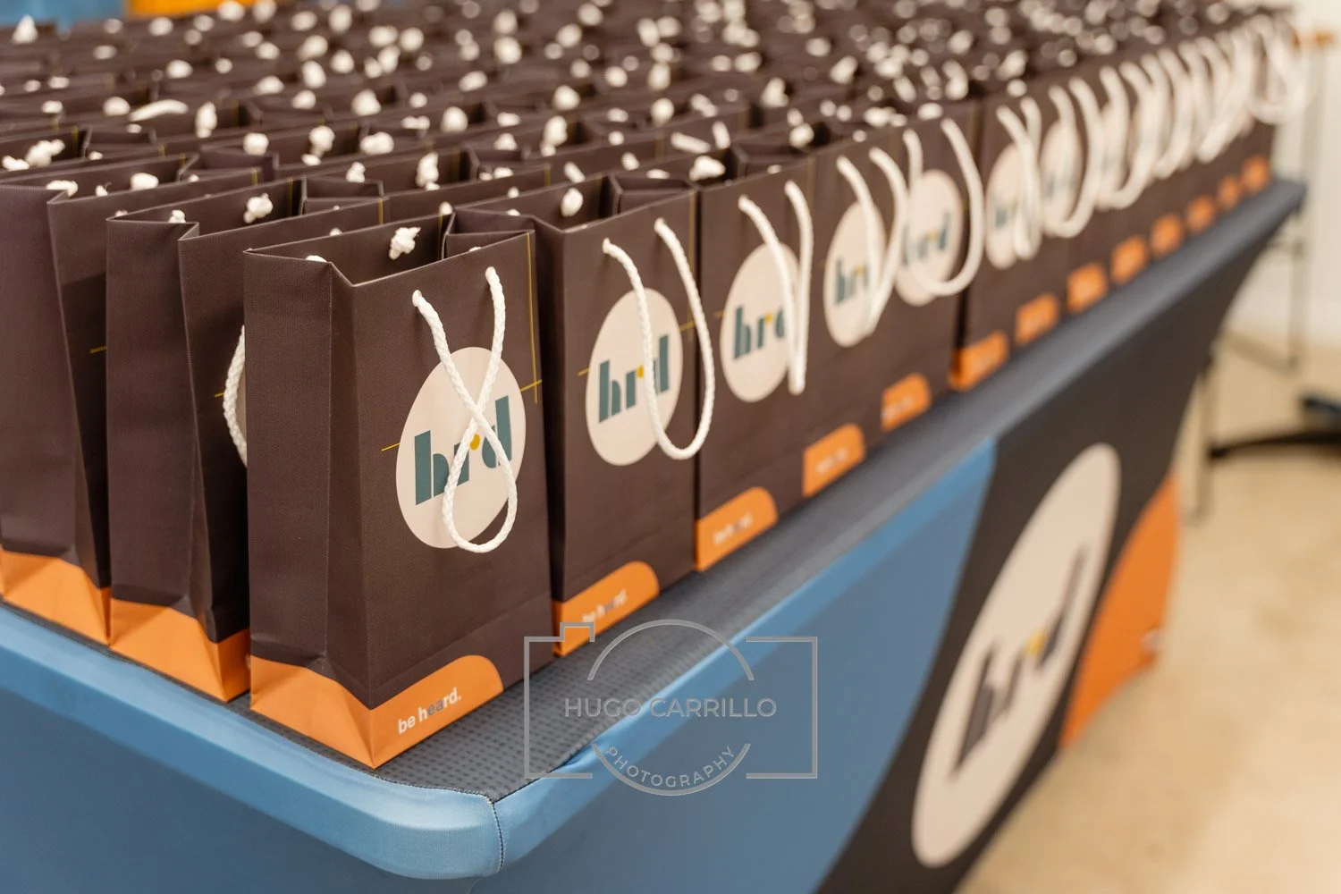 Multiple brown gift bags with white handles lined up on a table, each with a white circle logo and an orange label at the bottom that reads 'be heard.'