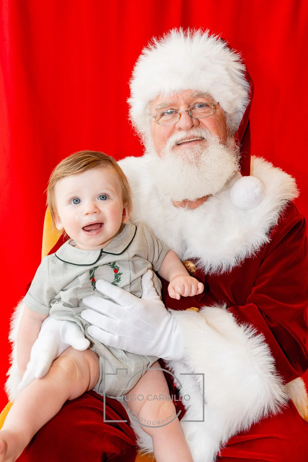 A young child sitting on Santa Claus's lap, both smiling, in front of a red backdrop, with Santa dressed in a traditional red and white costume with a Santa hat.