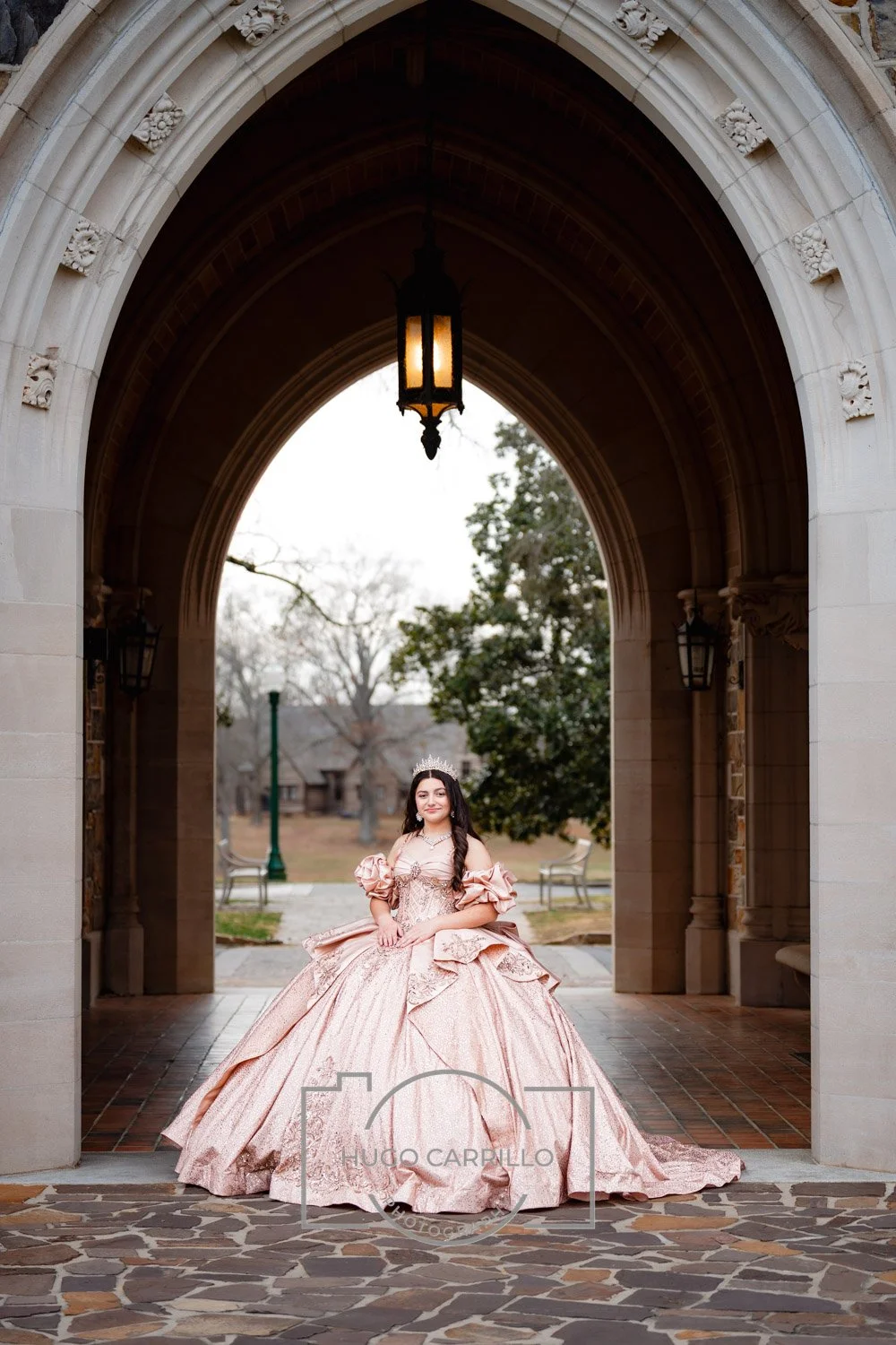 A quinceañera in an elaborate pink Victorian-style gown and tiara standing under an arched stone gateway with a lantern hanging above her.