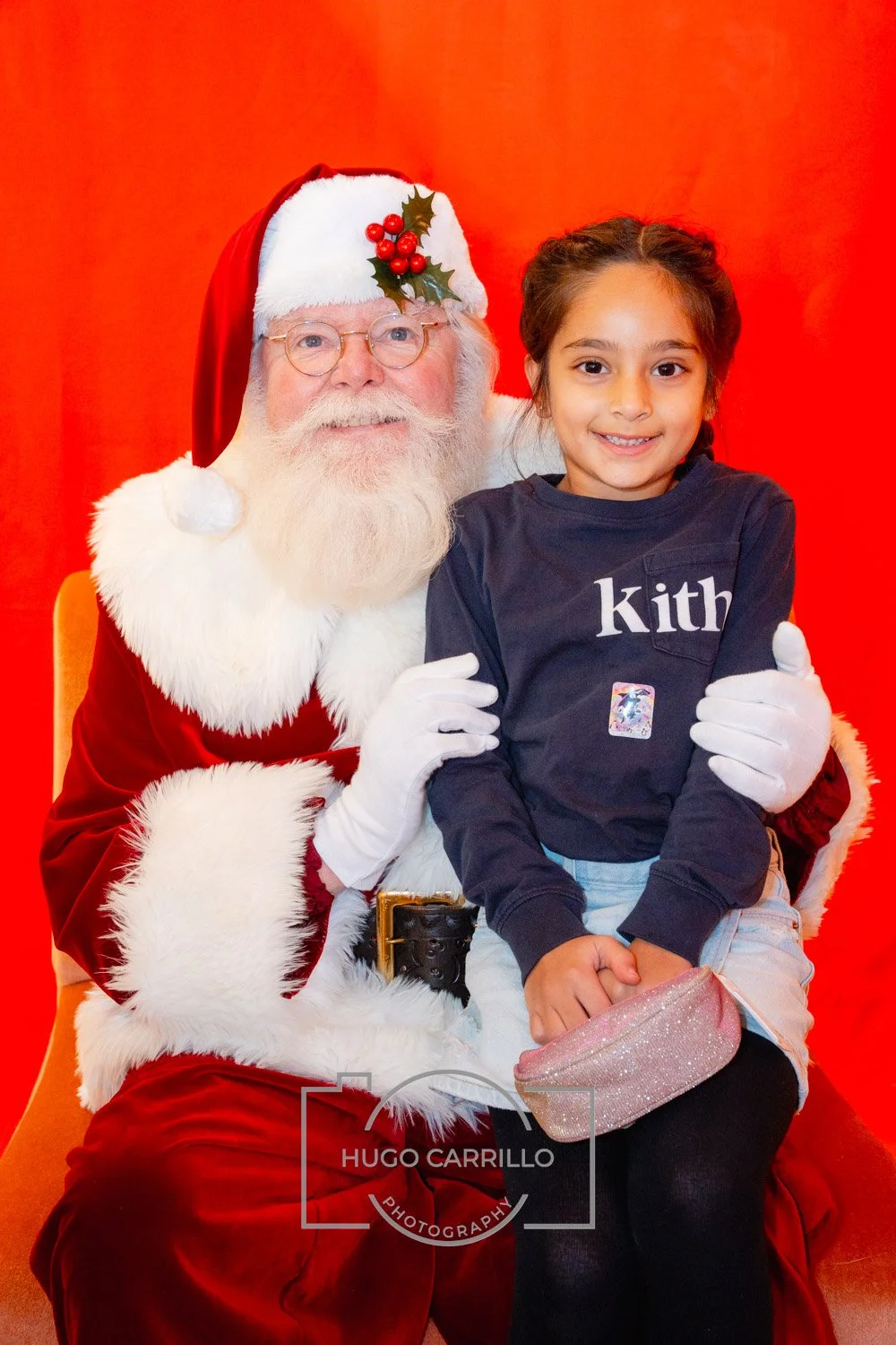 A young girl and a man dressed as Santa Claus sitting in front of a red background. The girl is smiling, wearing a navy blue sweatshirt with white text, holding a pink glittery purse, with her legs crossed. Santa Claus has a white beard, glasses, a r