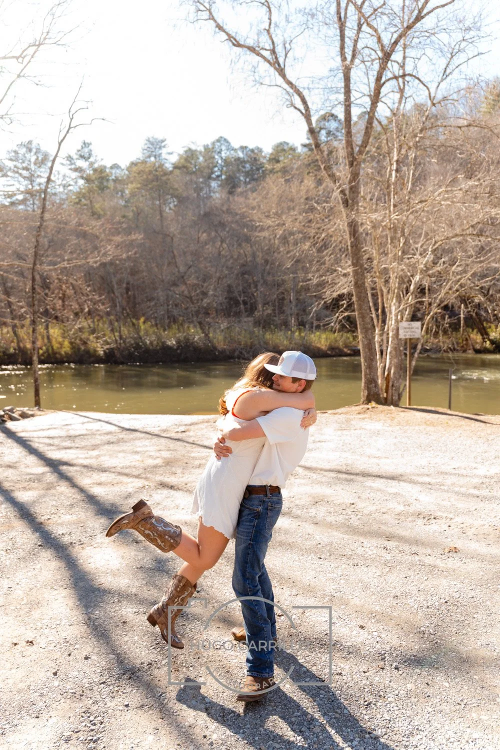 A couple embracing outdoors by a river on a sunny day, with the woman lifting her leg and wearing cowboy boots.