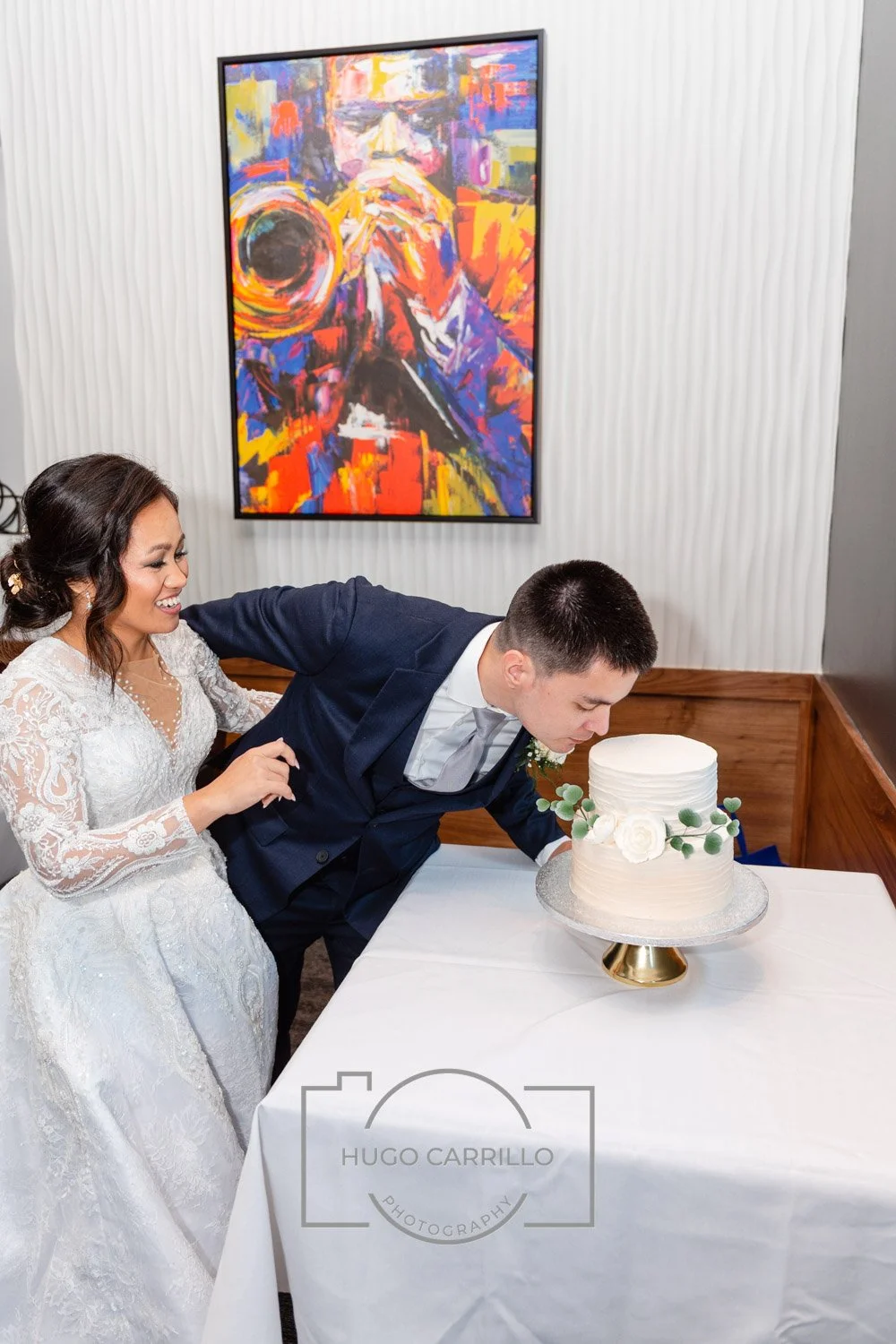 Couple at wedding cutting a white wedding cake with floral decorations, sitting at a table with a white tablecloth, colorful abstract painting on the wall.