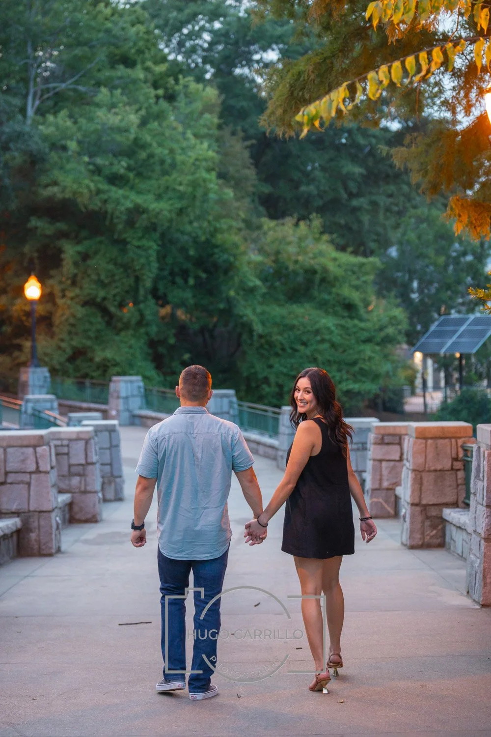 A couple holding hands and walking in a park during sunset, surrounded by trees and stone railings.