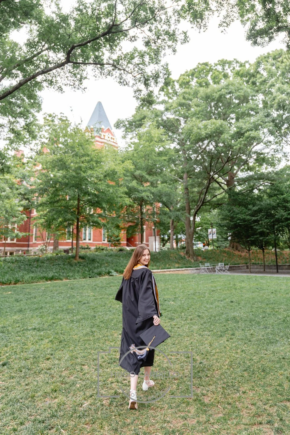 A young woman in graduation gown and cap walking on a grassy area in a park with trees and a brick building with a pointed roof in the background.