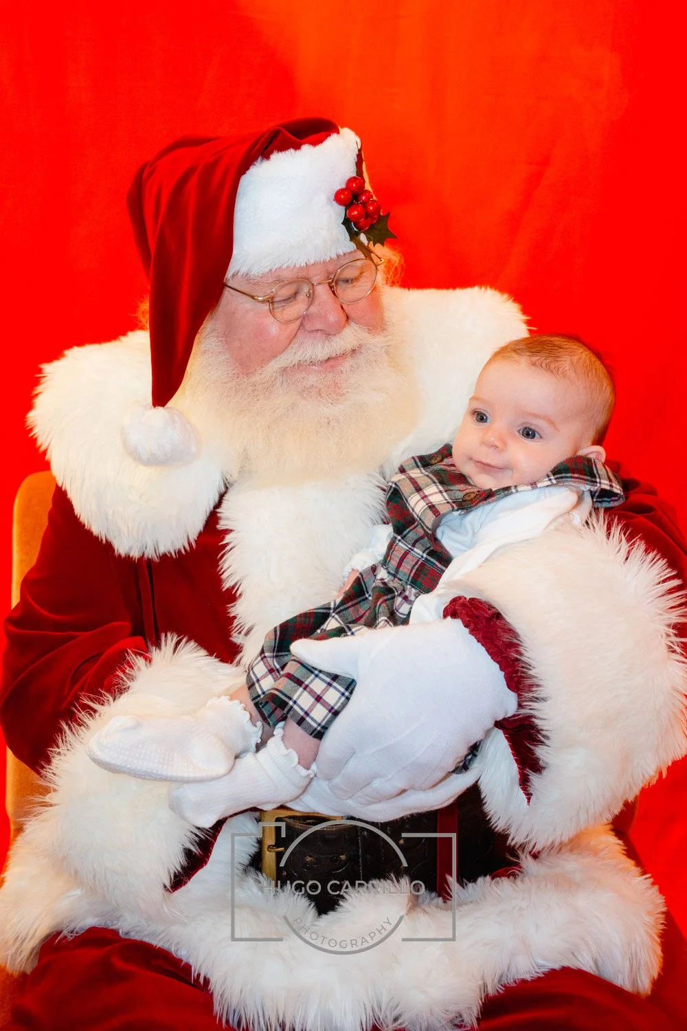 Santa Claus holding a baby at a holiday event, with a red background.