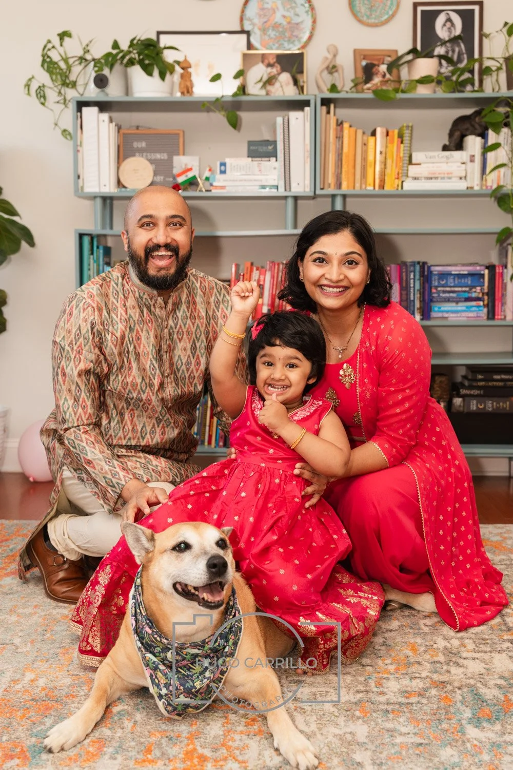 A happy family of three with a dog celebrating in their living room. The man has a beard and is wearing traditional Indian clothing, the woman has black hair and is wearing a red dress, and the girl is dressed in a matching red dress. They are smilin