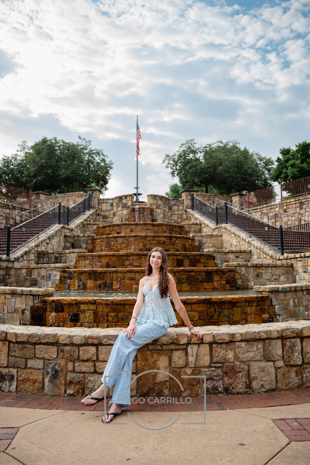 A young woman with long curly hair, wearing a light blue sundress and black sandals, sitting on a stone ledge in front of a tiered stone fountain with water flowing, outdoors during daytime with a cloudy sky, greenery, and an American flag in the bac