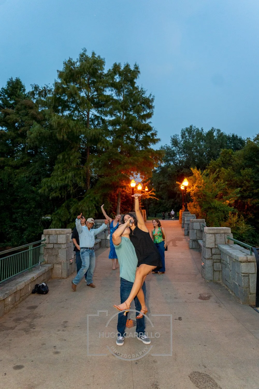 A group of people dancing and celebrating on a bridge at dusk with trees and street lamps in the background.