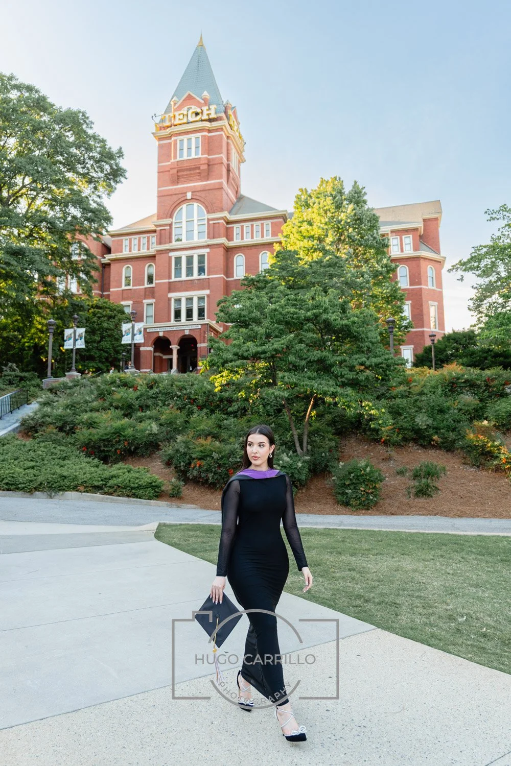 A woman in a black dress and high heels walking on a pathway in front of a large red brick building with a tall spire, surrounded by lush green trees and bushes.