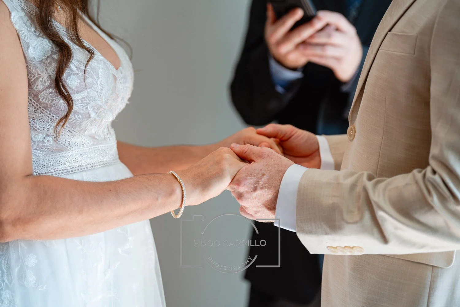 A couple holding hands during a wedding ceremony with an officiant in the background.