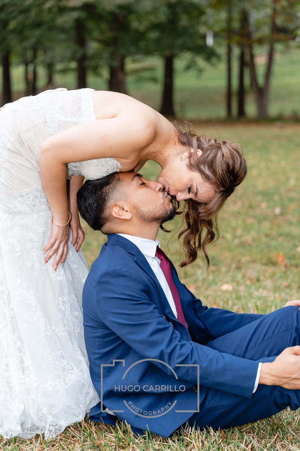 A bride and groom sharing a kiss outdoors, with the bride bending over the seated groom, in a park setting with green trees in the background.