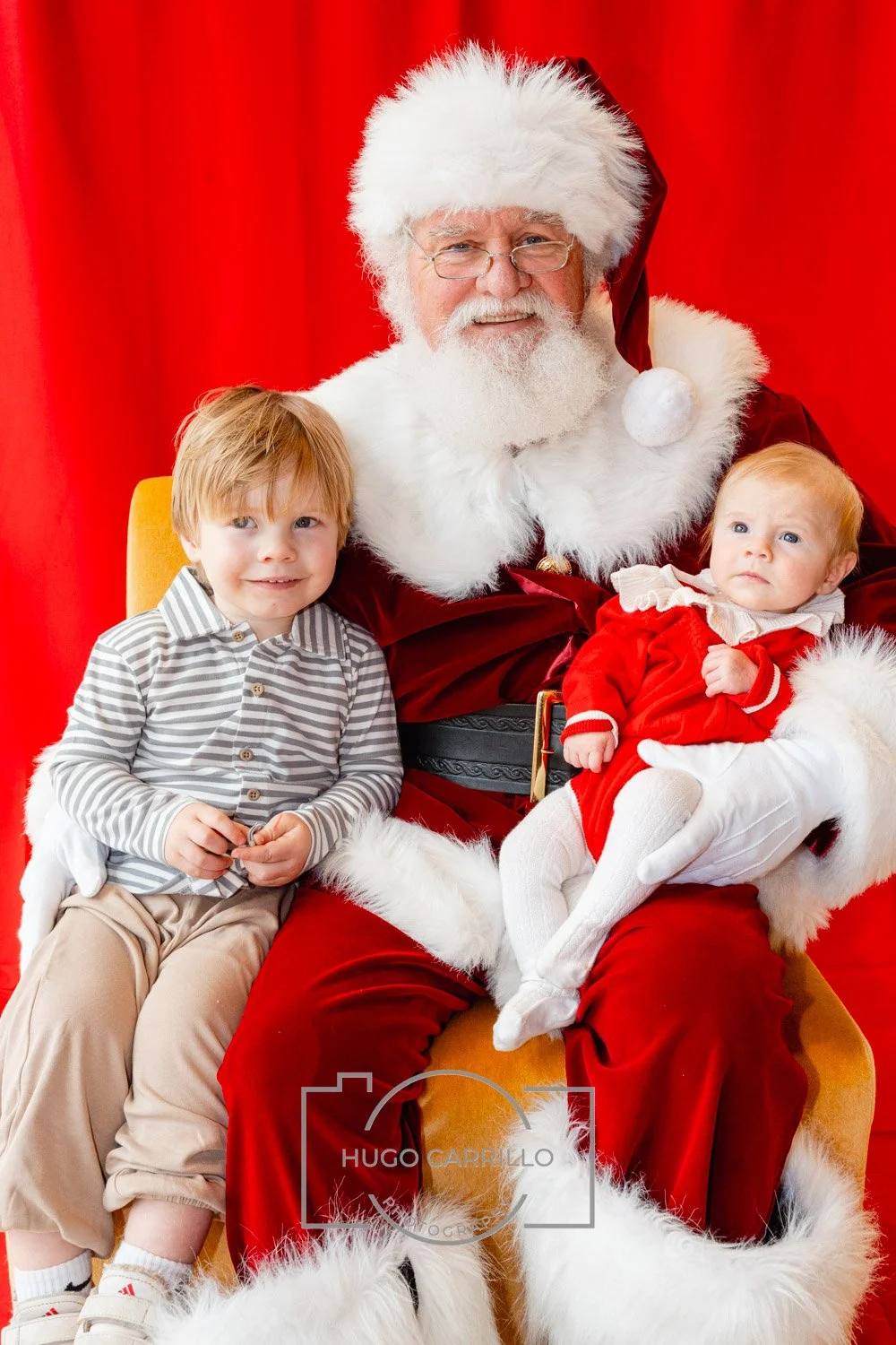 Santa Claus sitting on a yellow chair with a young boy on his left and a baby girl on his lap, all in front of a red curtain.