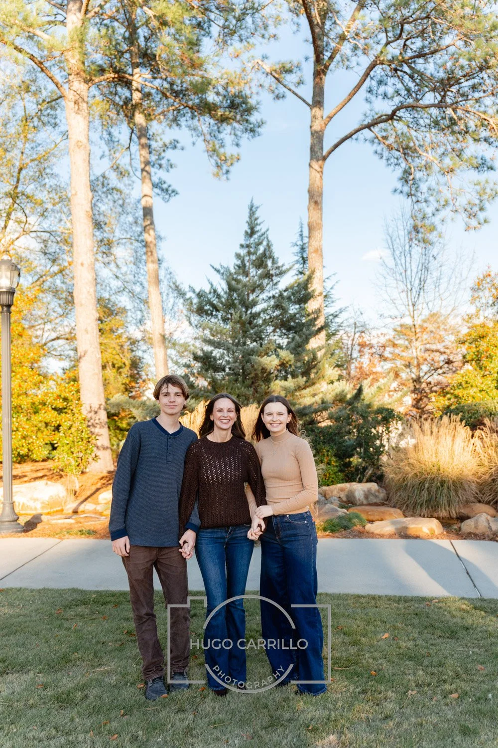 A woman with dark hair and two young adults, a man and a woman with brown hair, standing together outdoors in a park with trees and rocks, holding hands and smiling at the camera.