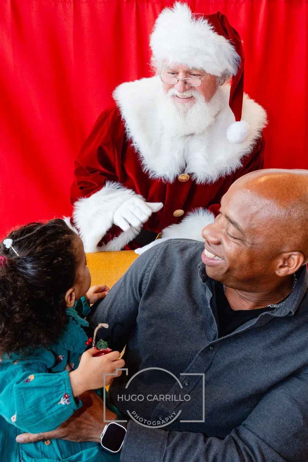 Santa Claus with glasses in a red and white outfit talking to a smiling man and a young girl in a festive setting.