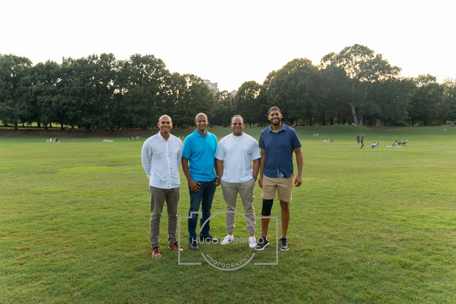 Four men standing on a grassy field in a park, smiling at the camera, with trees in the background and people relaxing in the distance during sunset.