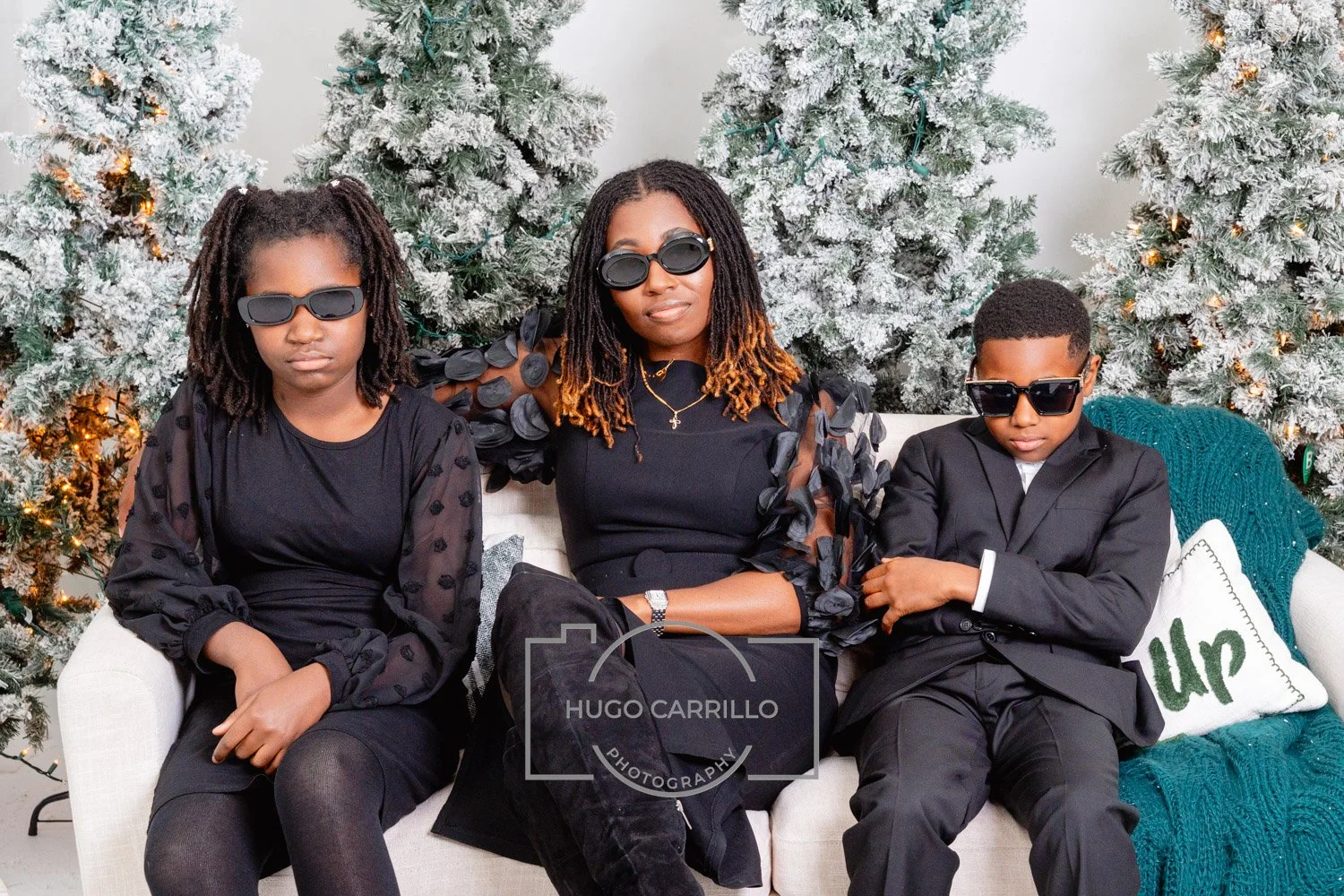 A woman and two children sitting on a white couch with snow-covered Christmas trees in the background, dressed in black and wearing sunglasses, with a knitted throw blanket and a pillow that says 'love'.