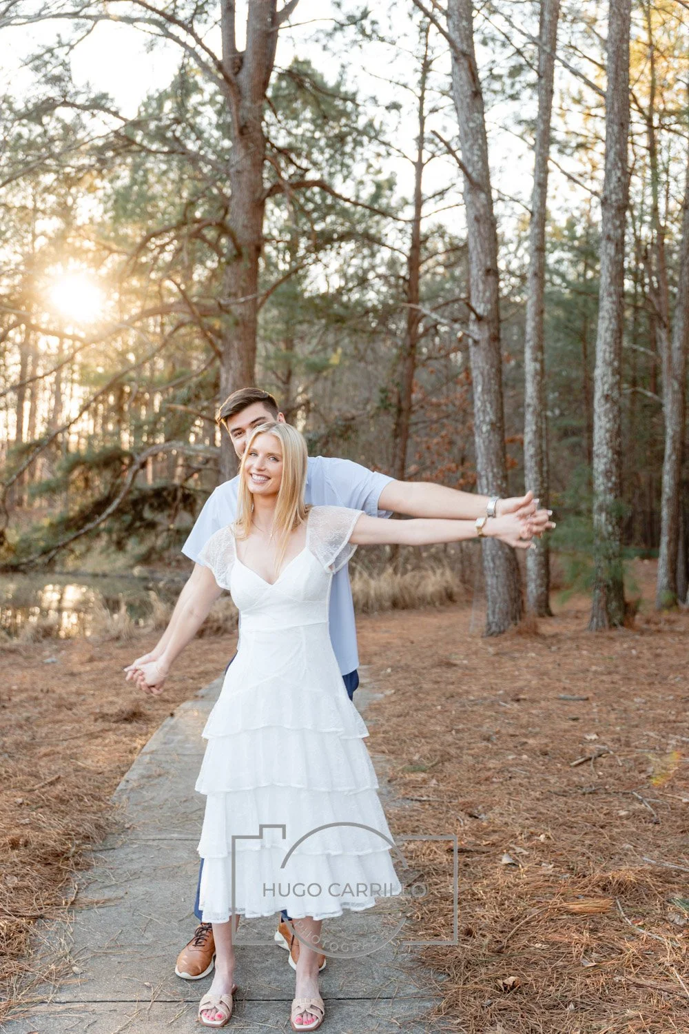A young couple standing on a wooden path in a forest during sunset, smiling and embracing each other, with tall trees and sunlight in the background.