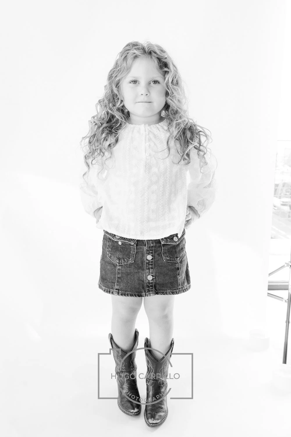 A young girl with curly hair standing with her hands behind her back, wearing a white blouse, a denim skirt, and cowboy boots, against a white background.