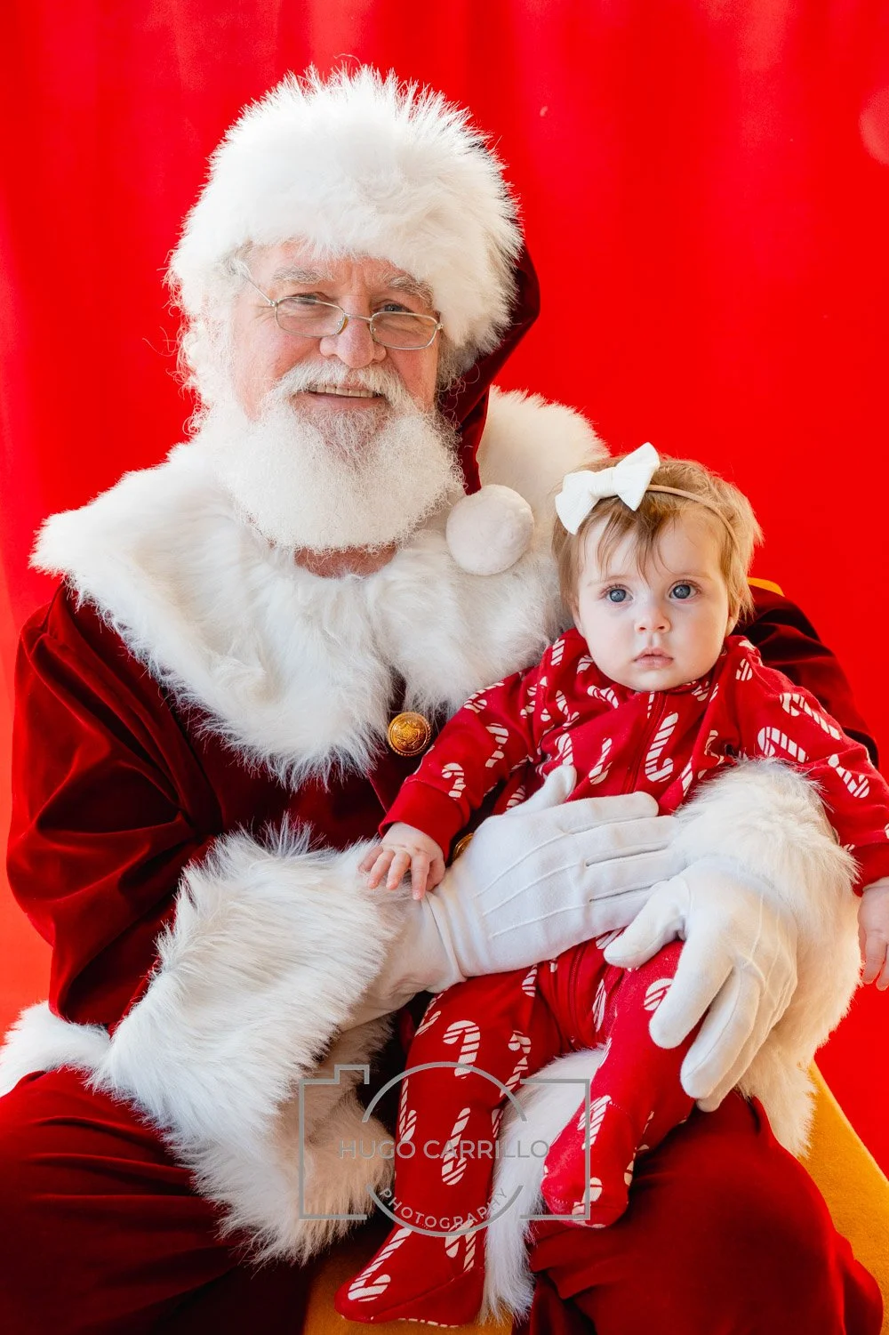 Santa Claus in a red velvet suit with white fur trim holding a young girl in Christmas pajamas against a red background.