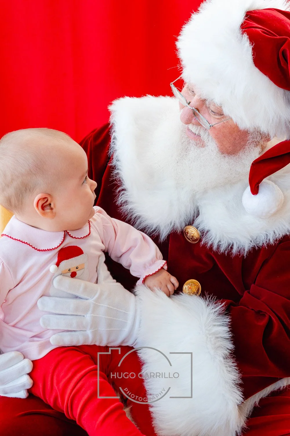 A young child sitting on Santa Claus's lap, looking into his eyes. Santa is wearing his traditional red suit with white fur trim and a red Santa hat. The child is dressed in pink pajamas with a Santa face on the front, and white gloves. The backgroun