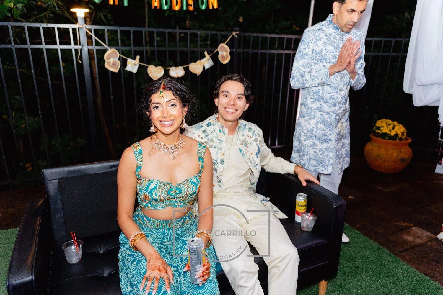 A couple dressed in traditional Indian attire sitting on a black couch, smiling at a celebration. The woman is wearing a blue patterned lehenga with jewelry, and the man is in a cream-colored sherwani with embroidery. Behind them, a man praying with 