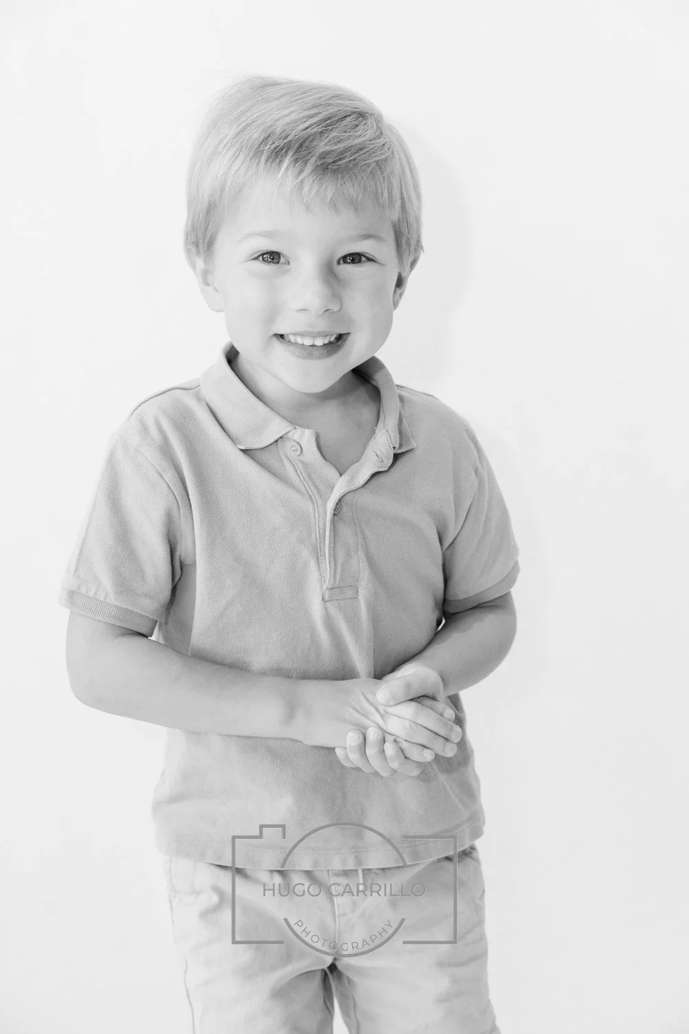 Black and white photo of smiling young boy with short blond hair, wearing a polo shirt and pants, standing against a plain light background.