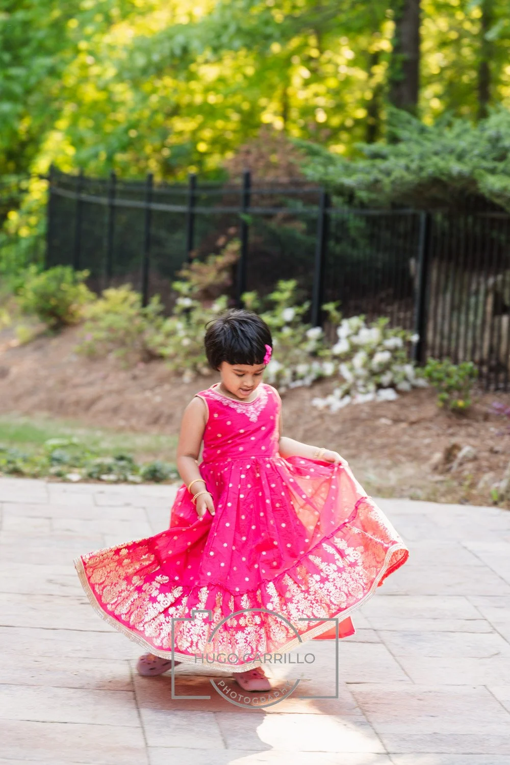 A young girl in a vibrant pink dress with gold accents twirling outdoors on a paved surface, with greenery and a black fence in the background.