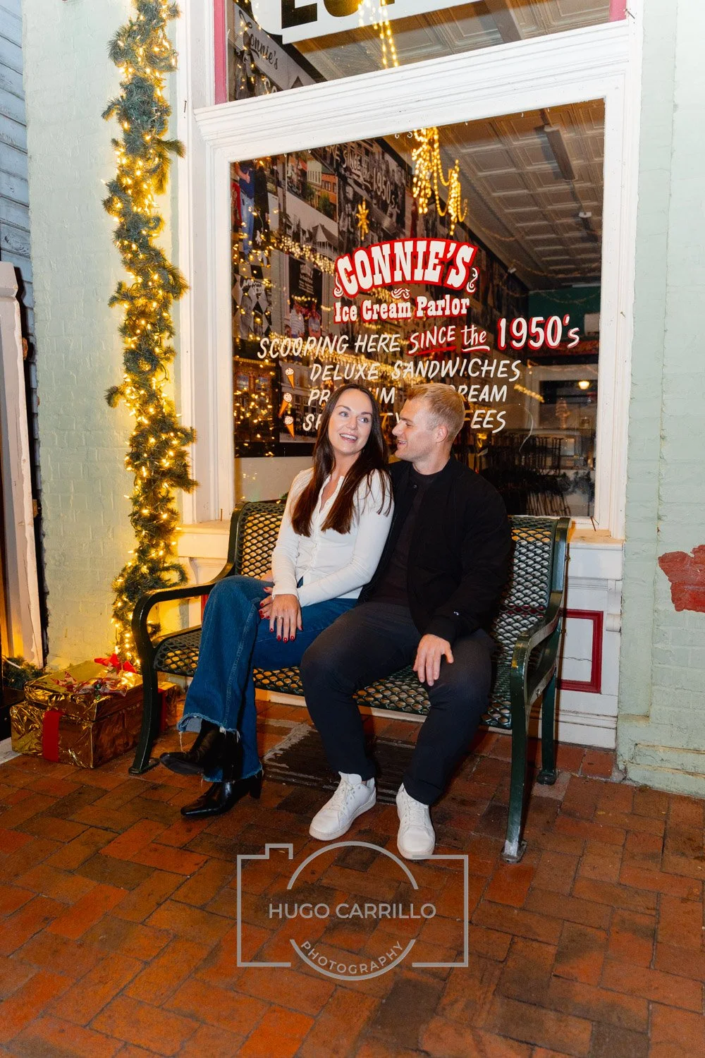 A young couple sitting on a black metal bench outside a vintage-style ice cream parlor decorated for Christmas with a garland of lights. The woman has long brown hair, white blouse, and blue jeans, and is smiling at the man. The man has short blond h