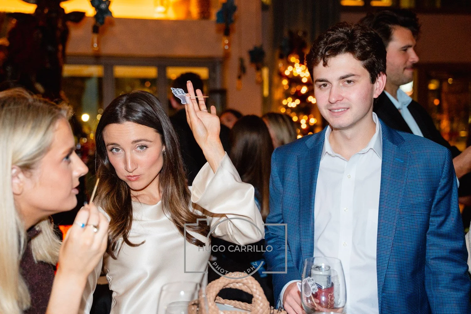 Group of people socializing at an indoor event with Christmas decorations, including a woman holding a cocktail pick with an appetizer, a woman in a cream satin blouse, and a man in a blue blazer holding a drink.