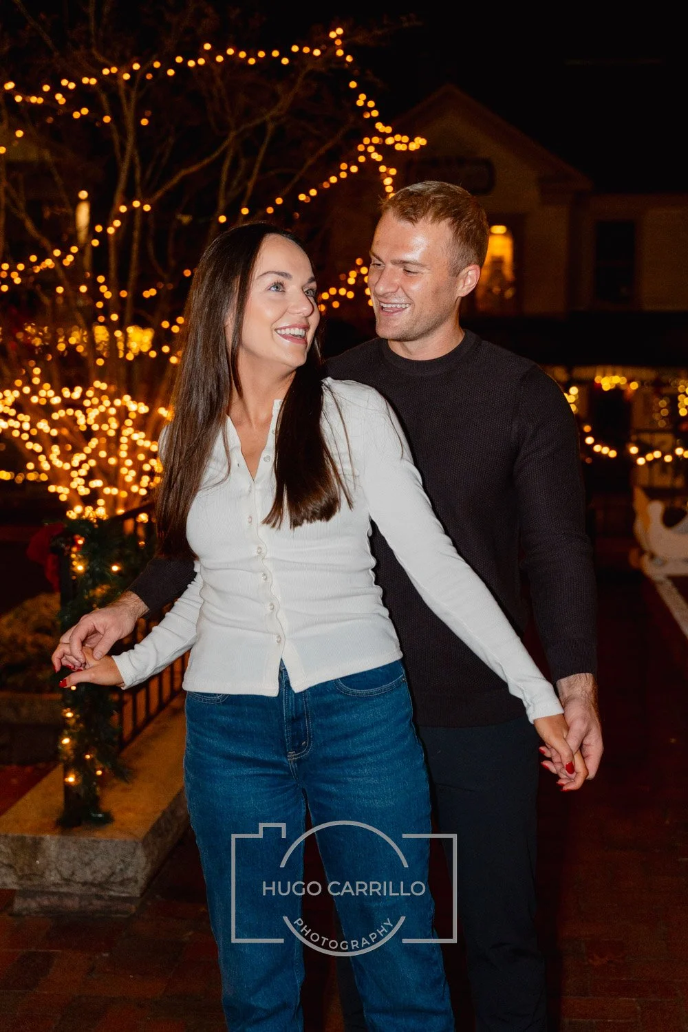 A happy couple holding hands and smiling at each other during night time with holiday lights in the background.