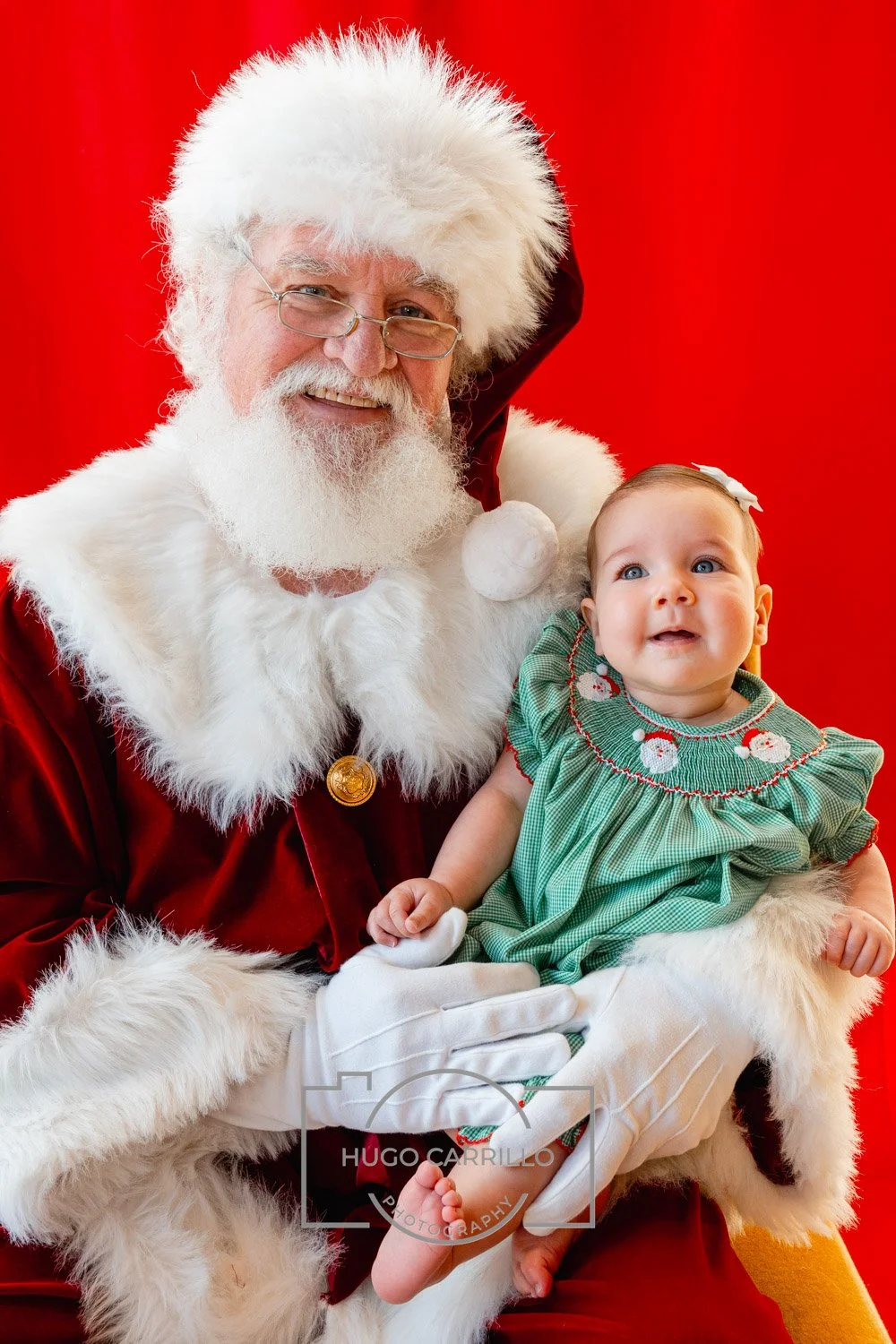Santa Claus, wearing glasses and a red suit with white fur trim, is smiling, holding a young girl dressed in a green Christmas dress with Santa embroidery, against a red background.