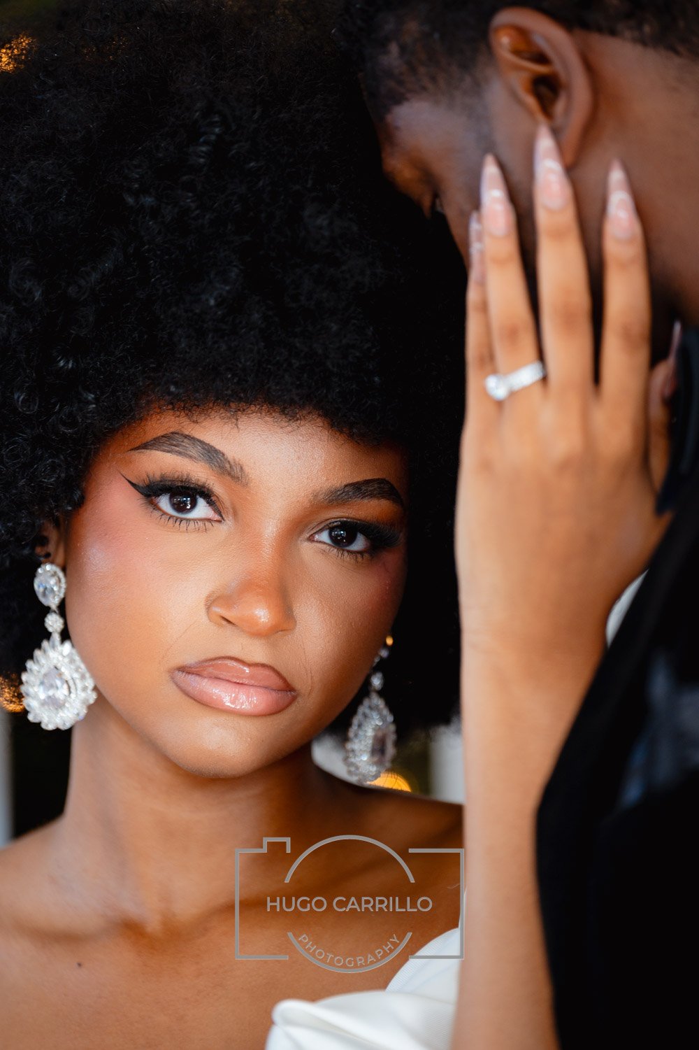 A woman with styled black hair and makeup looking into the camera, wearing large diamond earrings, and a ring on her finger, with a man showing his face partially and holding her face close. They appear to be in an intimate moment.