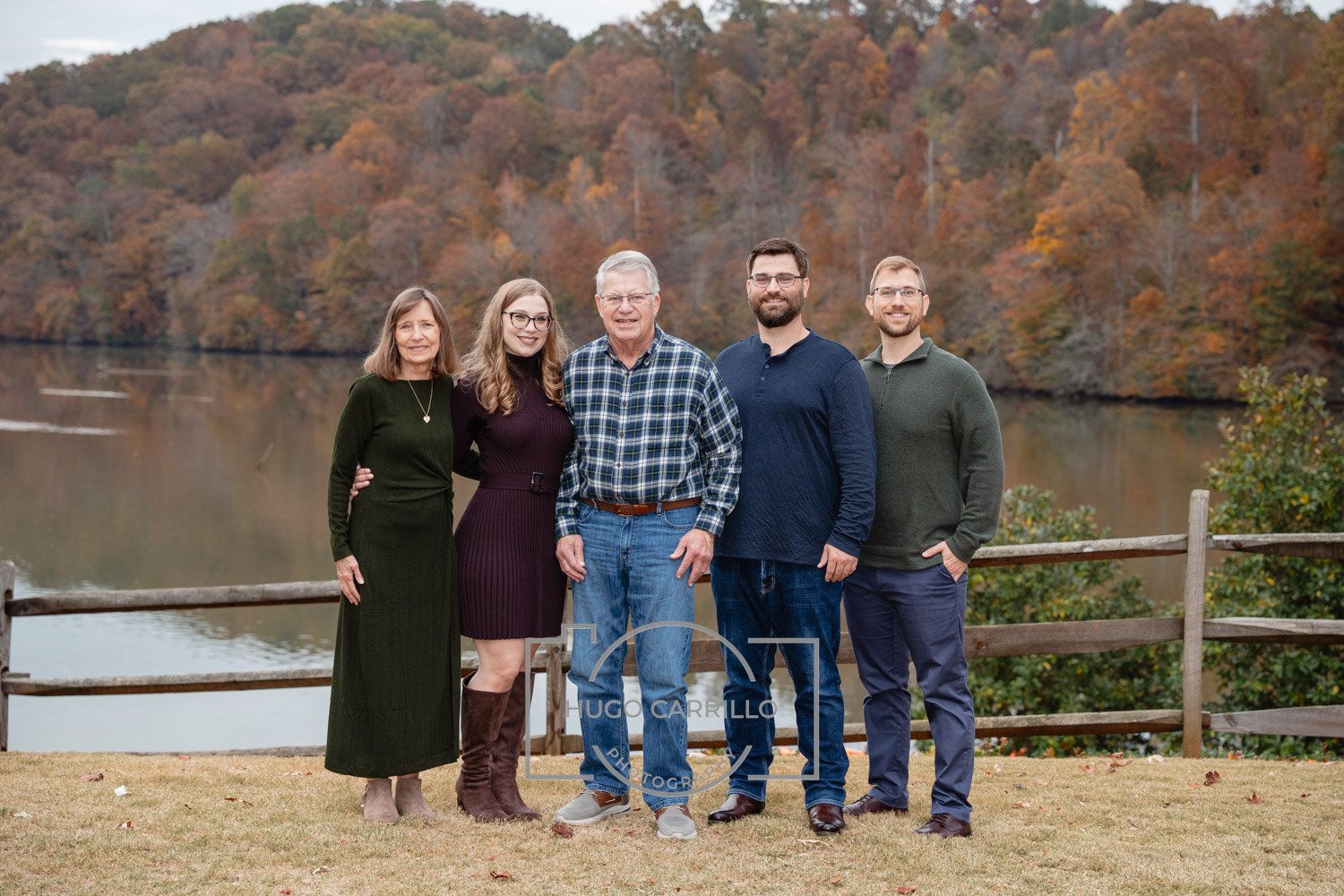 Family of five standing outdoors by a lake with fall foliage in the background.