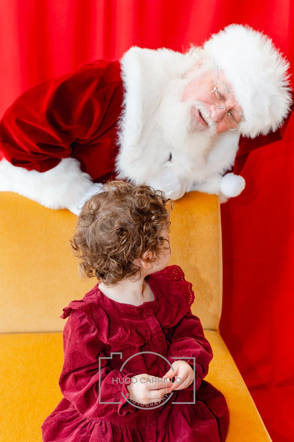 A young girl with curly hair wearing a red dress, sitting on a yellow chair, looking up at Santa Claus. Santa is leaning over and smiling, dressed in a red suit with white fur trim, against a red curtain background.