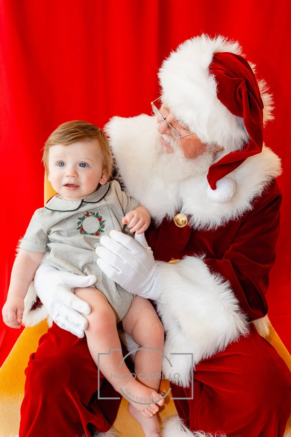 Santa Claus holding a young child on his lap, against a red backdrop, both looking at each other. Santa is dressed in traditional red and white with a red hat and glasses, and has a white beard. The child wears a gray shirt with a holiday wreath desi