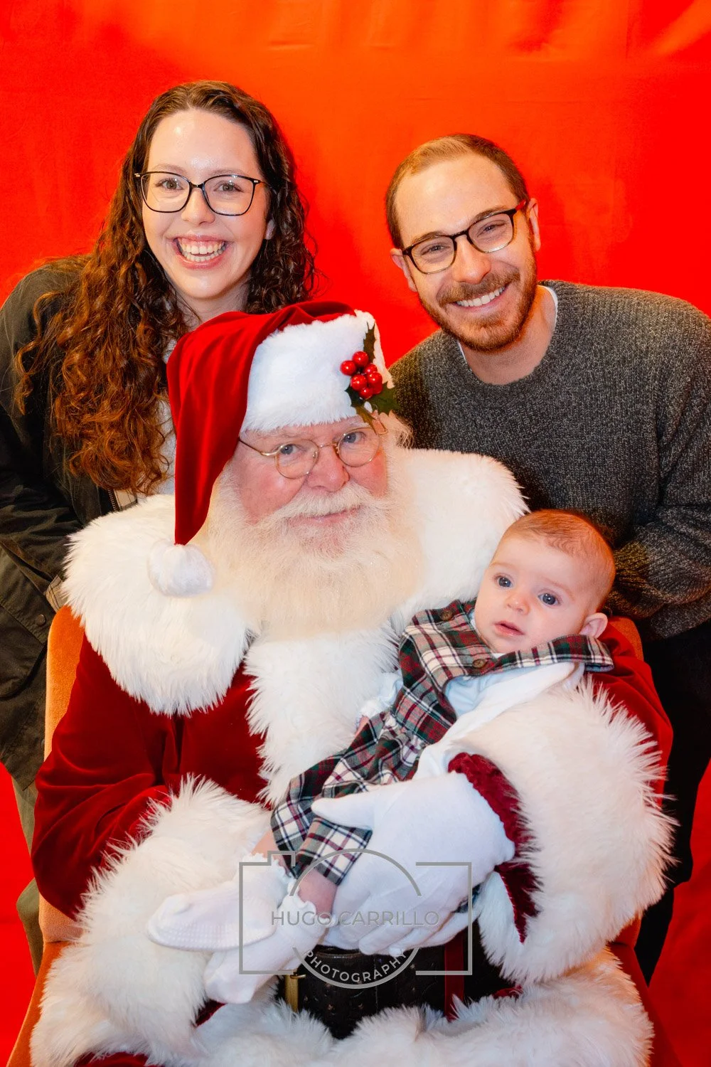 A family photo with Santa Claus. Santa is seated, holding a baby. Two adults stand behind them, smiling. The background is red.