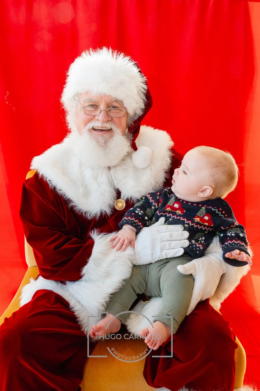 Santa Claus dressed in red and white, smiling, holding a young child, sitting against a red backdrop.