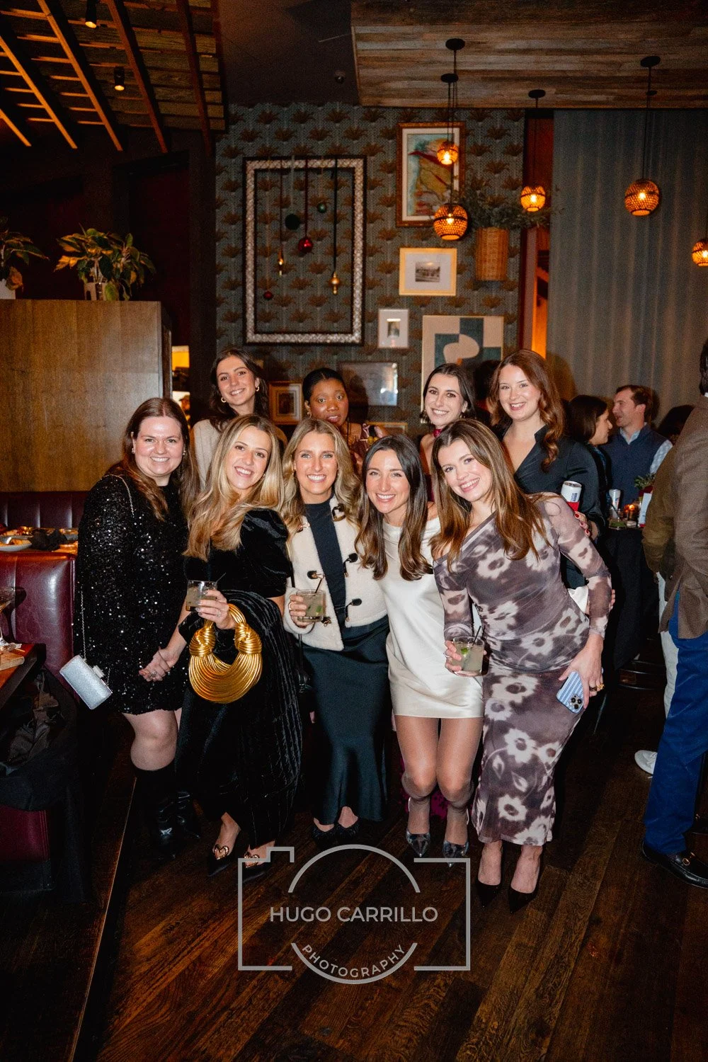A group of women celebrating at a party in a warmly decorated restaurant, smiling and holding drinks.
