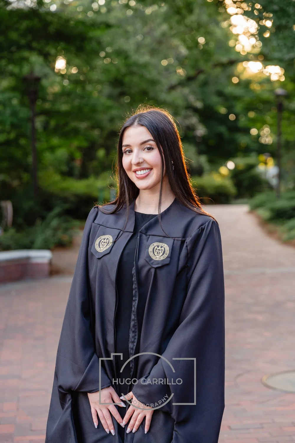 Young woman in a graduation gown standing outdoors on a brick path at sunset, smiling at the camera.