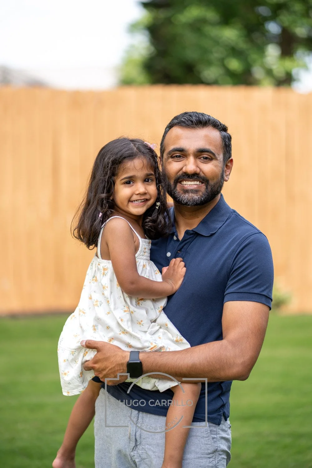 A man holding a young girl in his arms, both smiling at the camera in a backyard with a wooden fence and green trees in the background.