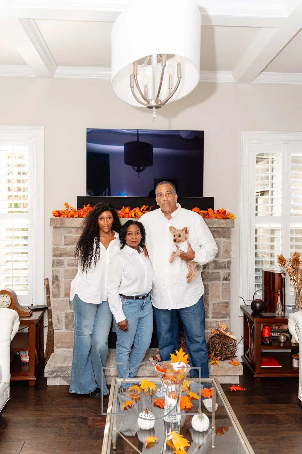 Family of four, two women, one man, and a small dog, standing in a living room with fall decorations, a fireplace, and a flat-screen TV behind them.