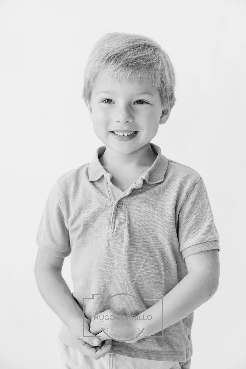 A young boy with light-colored hair, smiling, wearing a polo shirt, standing against a plain background.