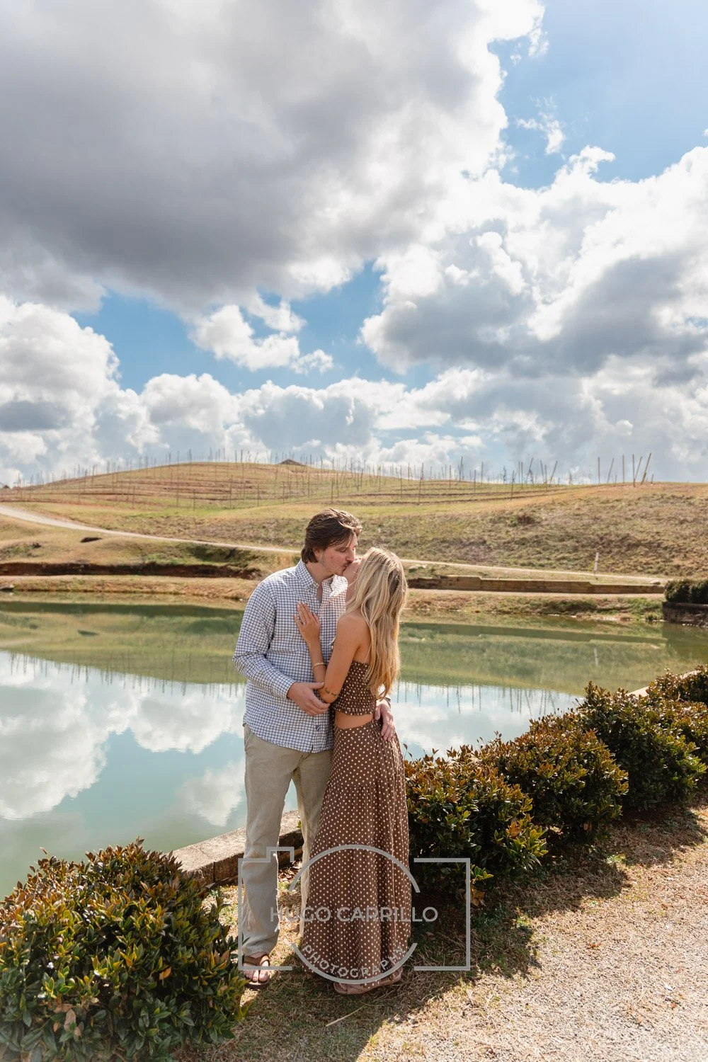 A couple standing by a pond on a cloudy day, sharing a kiss with hills in the background.