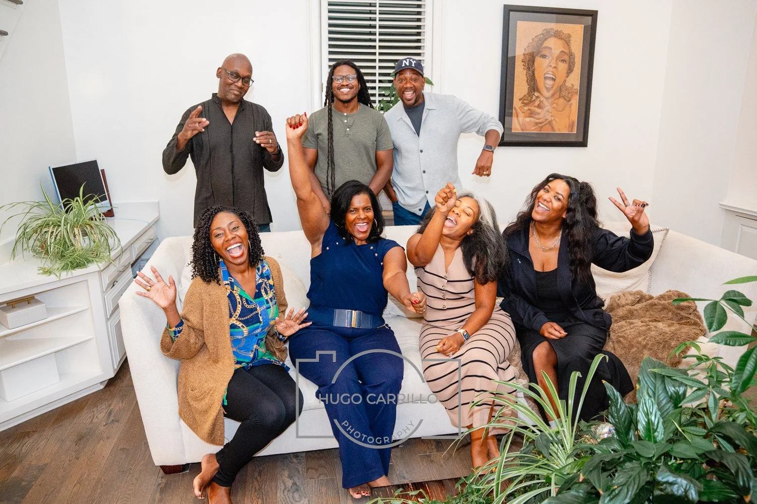 Group of eight diverse people sitting and standing on and around a white couch in a living room, smiling and celebrating. There are plants and framed art on the white walls, and a wooden floor.