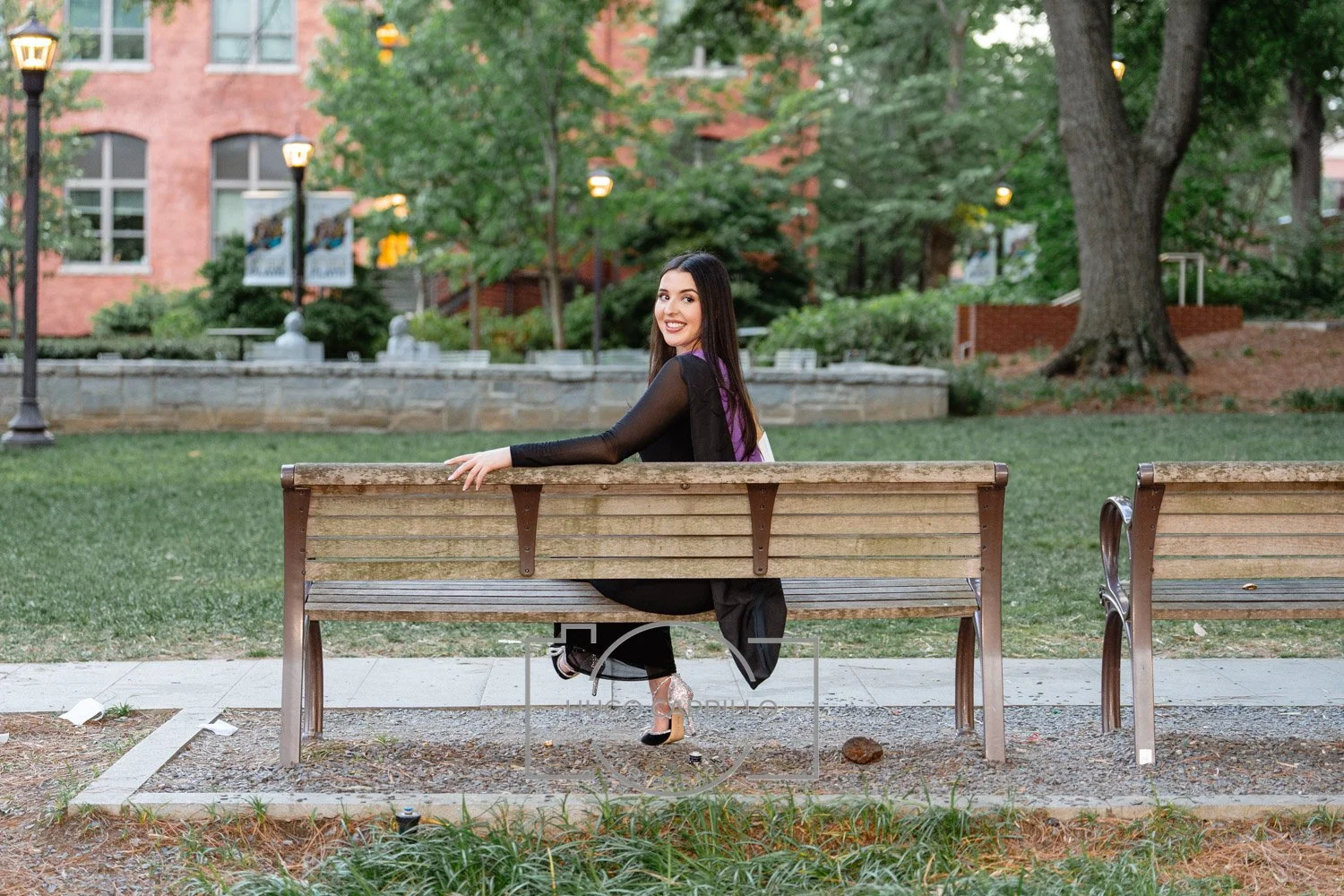 A woman with long dark hair, wearing a black dress with sheer sleeves, sitting on a park bench and smiling at the camera in a park with grass, trees, and a brick building in the background.