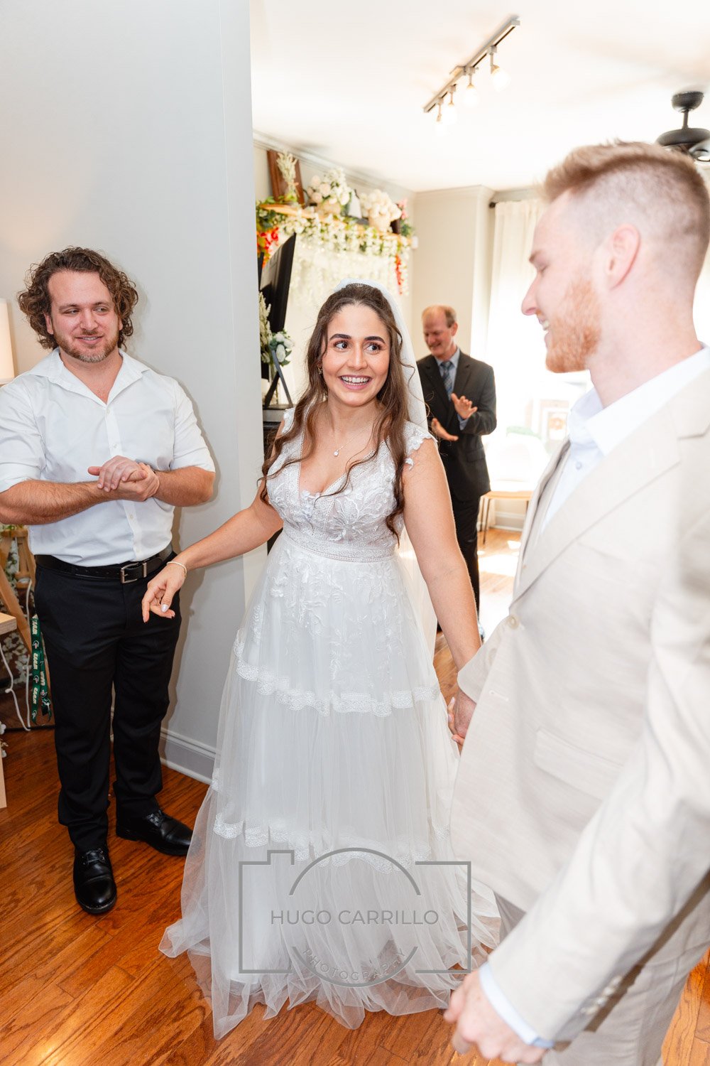 A bride in a white wedding dress holding hands with a groom in a white suit, smiling at each other, during a wedding celebration indoors, with two other men in the background.