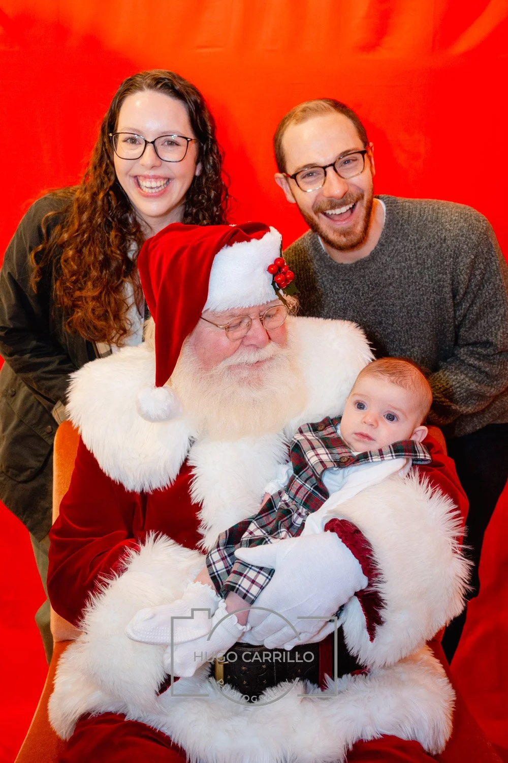 A group of four people, including a man dressed as Santa Claus holding a baby, a woman with glasses, and a man with glasses, smiling in front of a red background.