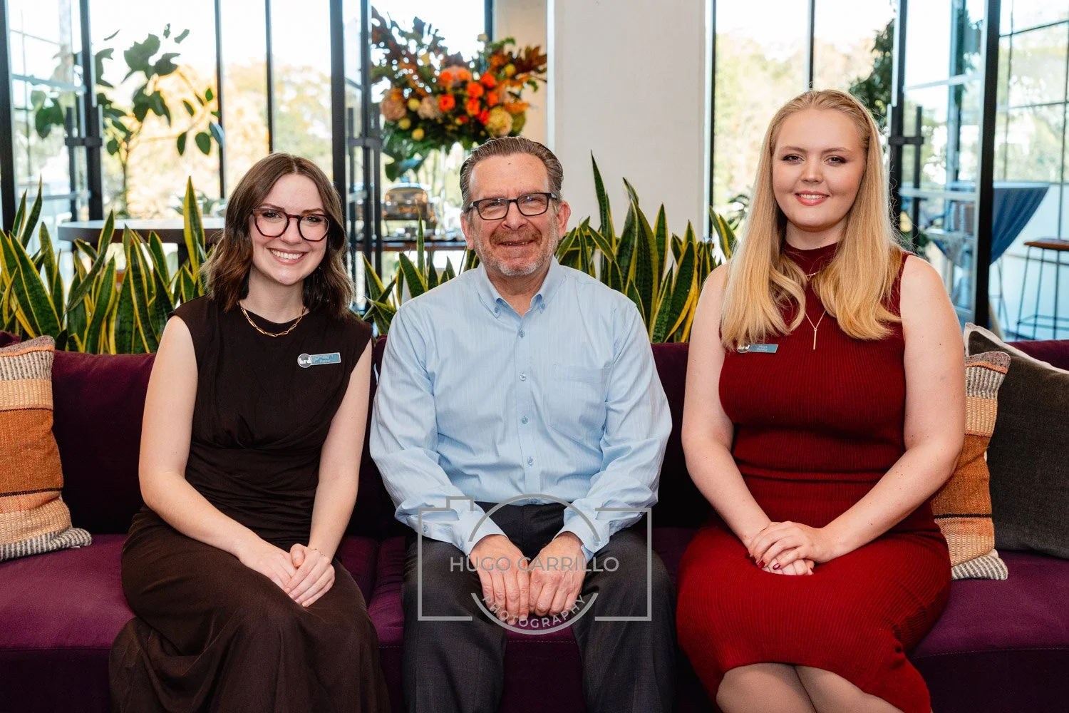 Three people sitting on a purple couch in a bright room with large windows and green plants. They are smiling and dressed professionally, with name tags visible.