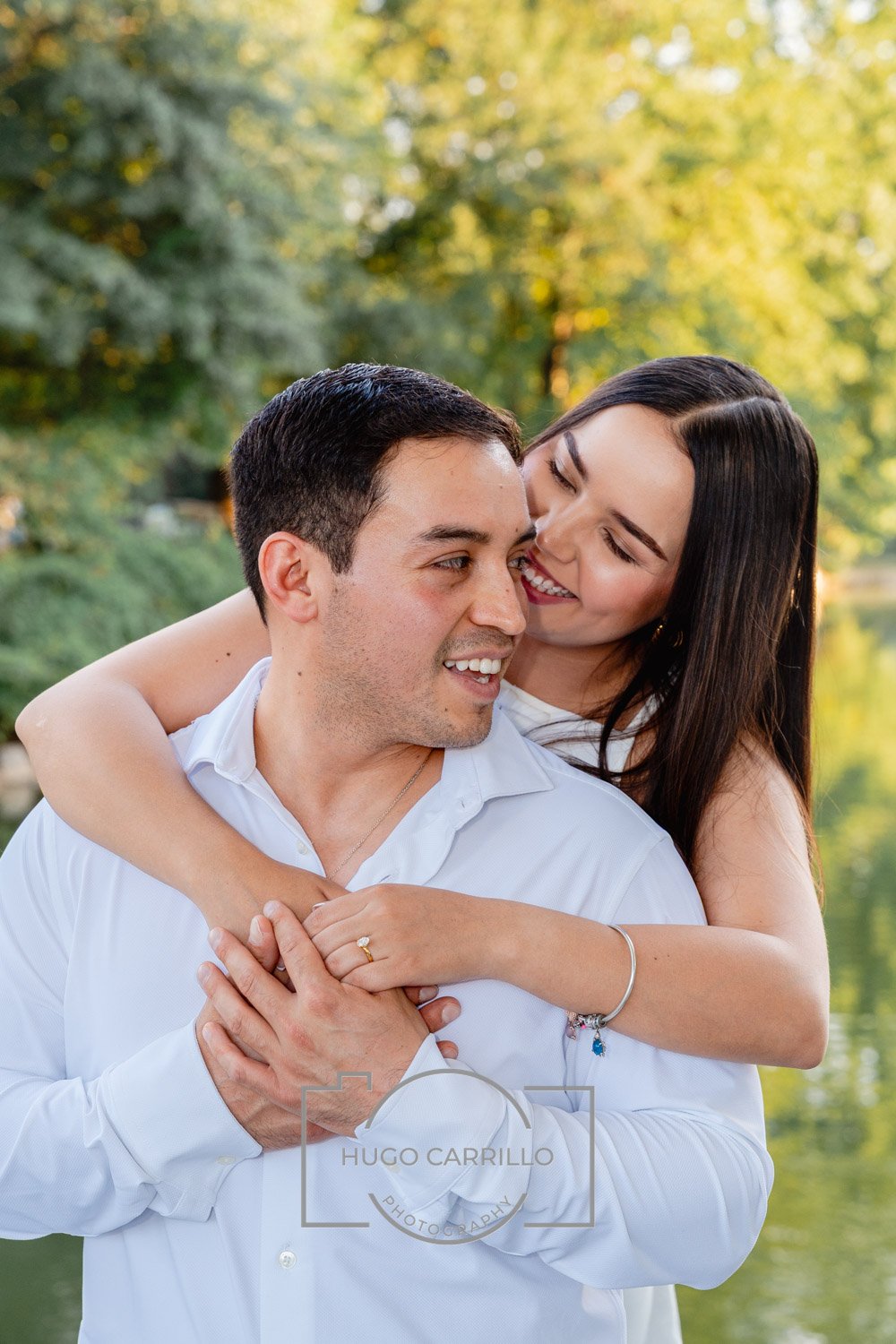 A couple smiling and embracing outdoors during daytime with a background of trees and sunlight.