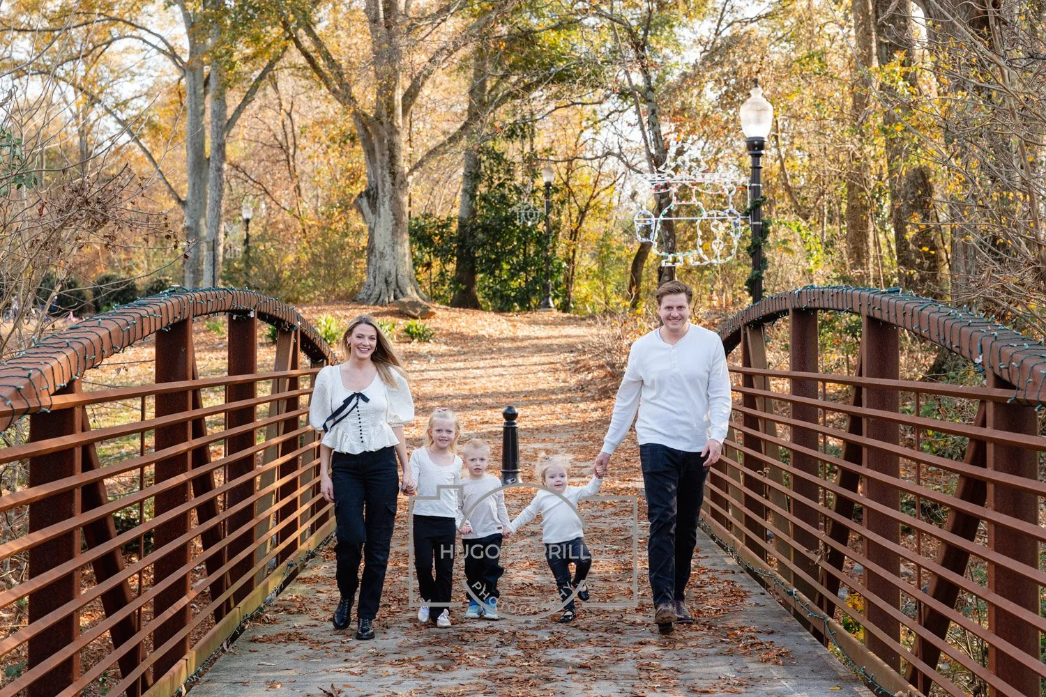 A family of six, including two adults and four children, walking across a wooden bridge in a park during autumn. The family is smiling and the background features trees with fall foliage and decorative holiday lights hanging from the trees.